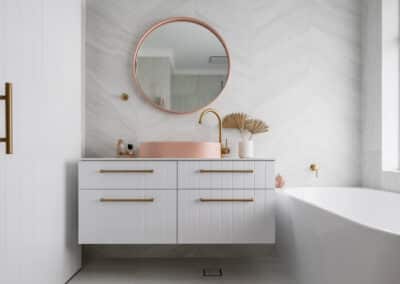 North beach coastal barn bathroom with brass tapware, pink concrete basins, v groove panelled cabinetry and onyx stone look tiles with a feature chevron pattern