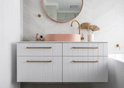 North beach coastal barn bathroom with brass tapware, pink concrete basins, v groove panelled cabinetry and onyx stone look tiles with a feature chevron pattern