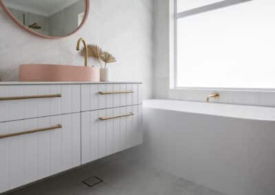 North beach coastal barn bathroom with brass tapware, pink concrete basins, v groove panelled cabinetry and onyx stone look tiles with a feature chevron pattern