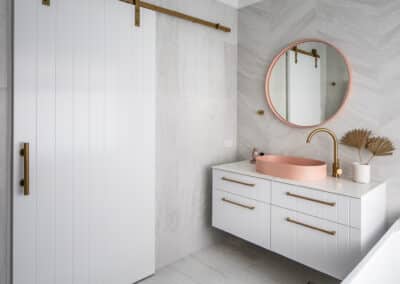 North Beach Coastal Barn Bathroom with brass tapware, pink concrete basins, V Groove panelled cabinetry and Onyx stone look tiles with a feature chevron pattern