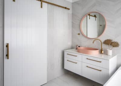 North beach coastal barn bathroom with brass tapware, pink concrete basins, v groove panelled cabinetry and onyx stone look tiles with a feature chevron pattern